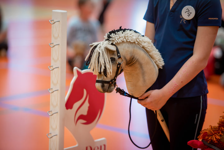 Közeli kép a LarDen és a CHHA fedett pályás versenyéről: sötétkék sportpólós résztvevők Hobby Horse lovakkal fehér díjugrató akadályoszlop és piros-fehér akadályszárny mellett; az előtérben egy drapp Hobby Horse ló krém-fekete fonalsörénnyel és kantárral.