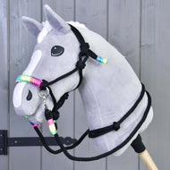 Fehér hókás szürke Hobby Horse ló a fekete csomózott kötőfékkel és kantárszárral, szivárványszínű tekeréssel, szürke fafal mellett