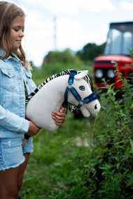 Girl in a denim jacket holding Hobby Horse Aurora in a blue halter outdoors by green plants, a red tractor behind