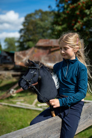Kékeszöld felsős szőke lány egy fakorláton ül, és a fekete kötőfékkel kapható Hobby Horse Bond lovat tartja, itt fekete kantárral és szárral