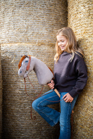 Smiling girl in a dark sweatshirt and jeans leaning on straw bales with Hobby Horse Chanell in a brown bridle and reins