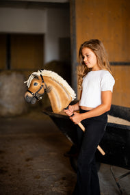 Fehér felsős lány áll az istállóban egy talicska mellett, kezében a fekete kötőfékes Hobby Horse Colby ló