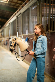 Farmerdzsekis, farmernadrágos lány egy istállófolyosón, a fekete kantárszárnál fogva tart egy fekete szőrmés kötőfékes, krémszínű Hobby Horse lovat
