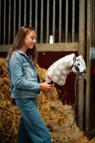 Girl in a denim jacket by straw bales holds Hobby Horse Elektra, here in a dark grey fur halter with a braided grey lead rope