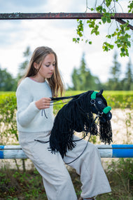 Fehér pulóveres lány egy akadályrúdon ül, és összefogja a zöld szőrmés kötőfékes fekete Hobby Horse ló kantárszárát