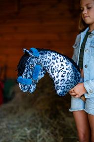 Girl in a denim jacket in a stable holds a blue and white leopard-print Hobby Horse wearing the petrol fur halter