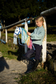 Mentazöld pulóveres lány a pálya fehér korlátja mellett, egy szivárványos sörényű fehér Hobby Horse lovat tart a kék csíkos vezetőszáron