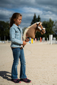 Farmerdzsekis lány egy akadályokkal berendezett homokos pályán, kezében világosbarna Hobby Horse ló a krémszínű szőrmés kötőfékben
