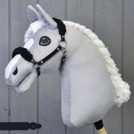 Szürke plüss Hobby Horse ló fonott fehér sörénnyel, rajta a fekete kötőfék fekete szőrmével, szürke fafal előtt