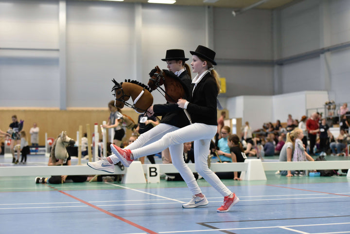 Two children in formal Hobby Horsing dress code outfits competing indoors with Hobby Horses