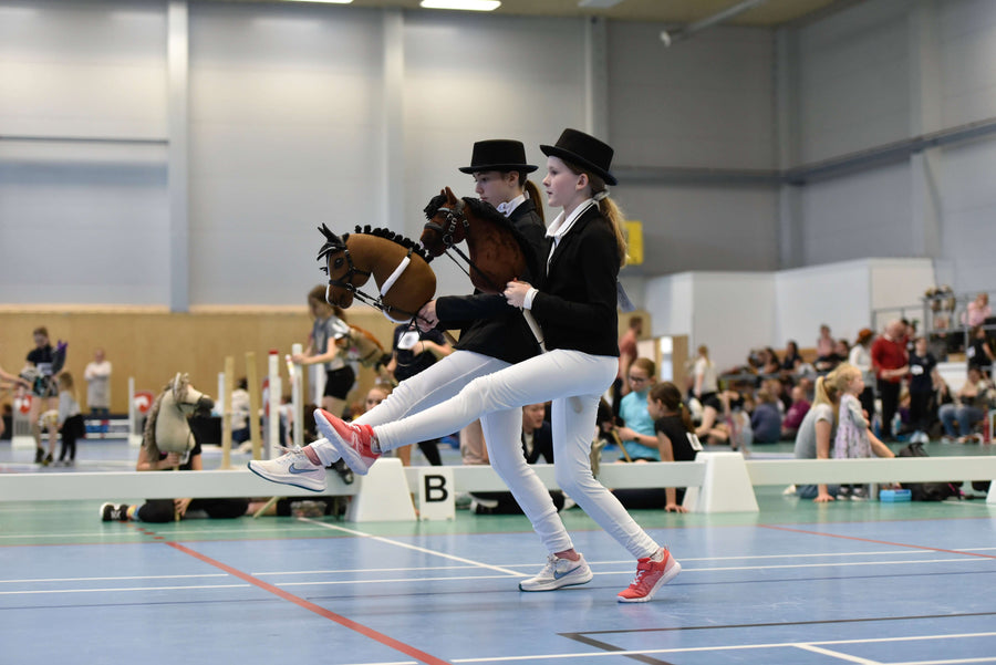 Két gyerek a Hobby Horsing öltözködési előírásainak megfelelő ünnepélyes ruhában versenyez Hobby Horse lovakkal egy teremben