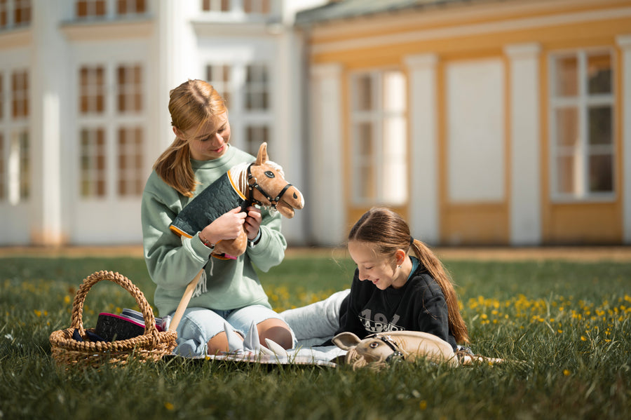 Halványzöld pulóveres lány ül a fűben egy felszereléssel teli fonott kosár mellett, és gyengéden tart egy drapp Hobby Horse lovat fekete kantárral, fehér sörénnyel és steppelt takaróval. A közeli pléden egy másik gyerek is részben látszik.