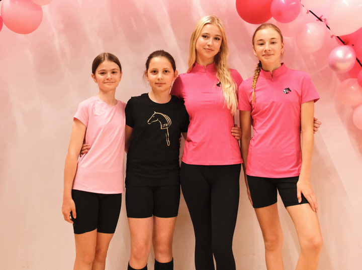 Négy fiatal sportoló áll egymásba karolva egy rózsaszín lufikkal díszített háttér előtt, sportos rózsaszín-fekete felsőben, fekete rövidnadrágban vagy leggingsben, Hobby Horse logós részletekkel. A vidám LarDen díszlet hívogató hangulatot áraszt.