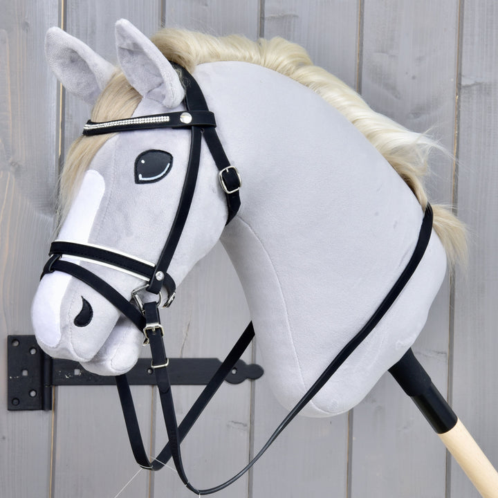 Plüss Hobby Horse ló fehér sörénnyel, fekete kantárral és fehér csíkkal az arcán, faboton, szürke fa háttér előtt.