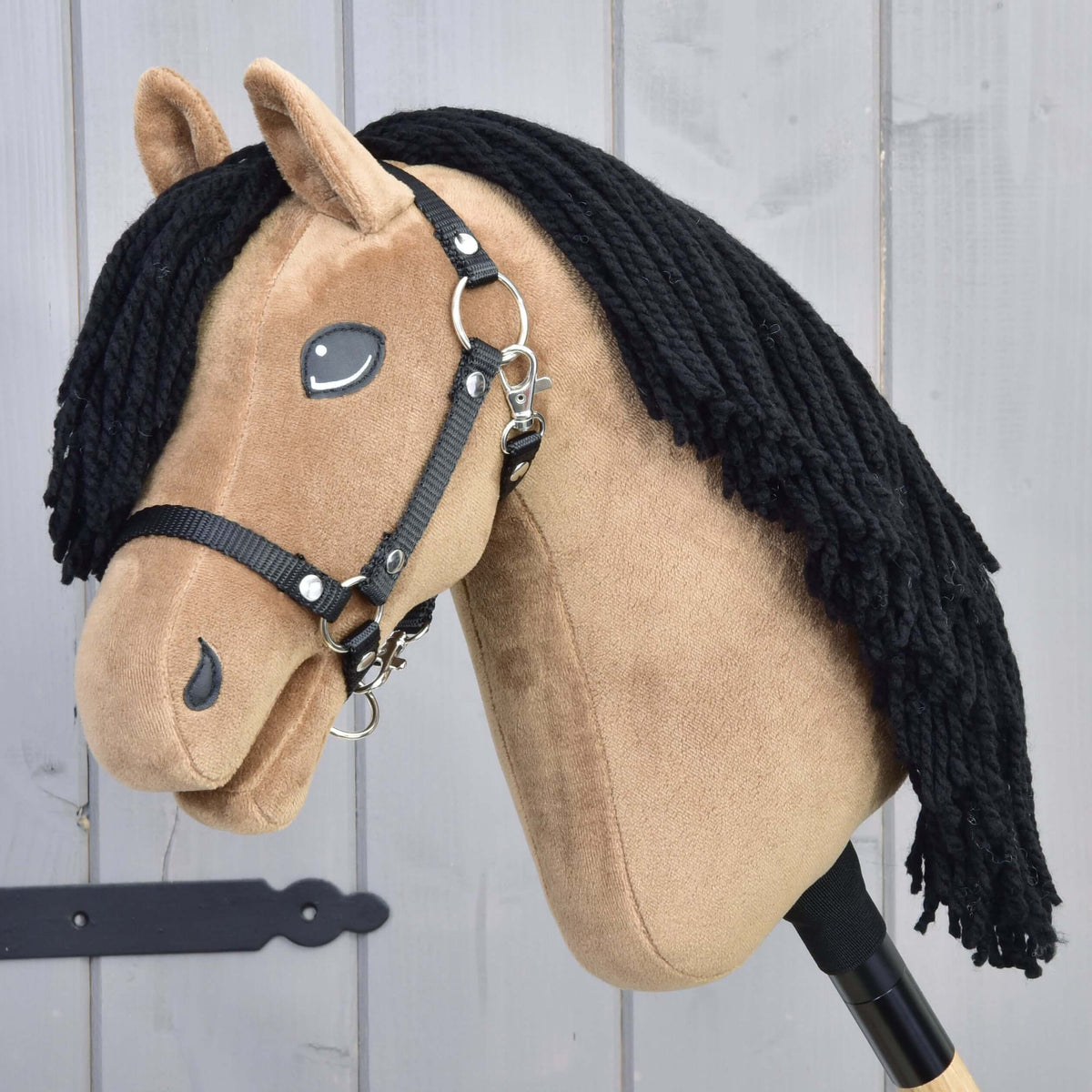 LarDen Hobby Horse Samantha with Black Halter, sandy plush head and long black yarn mane by a grey wooden wall