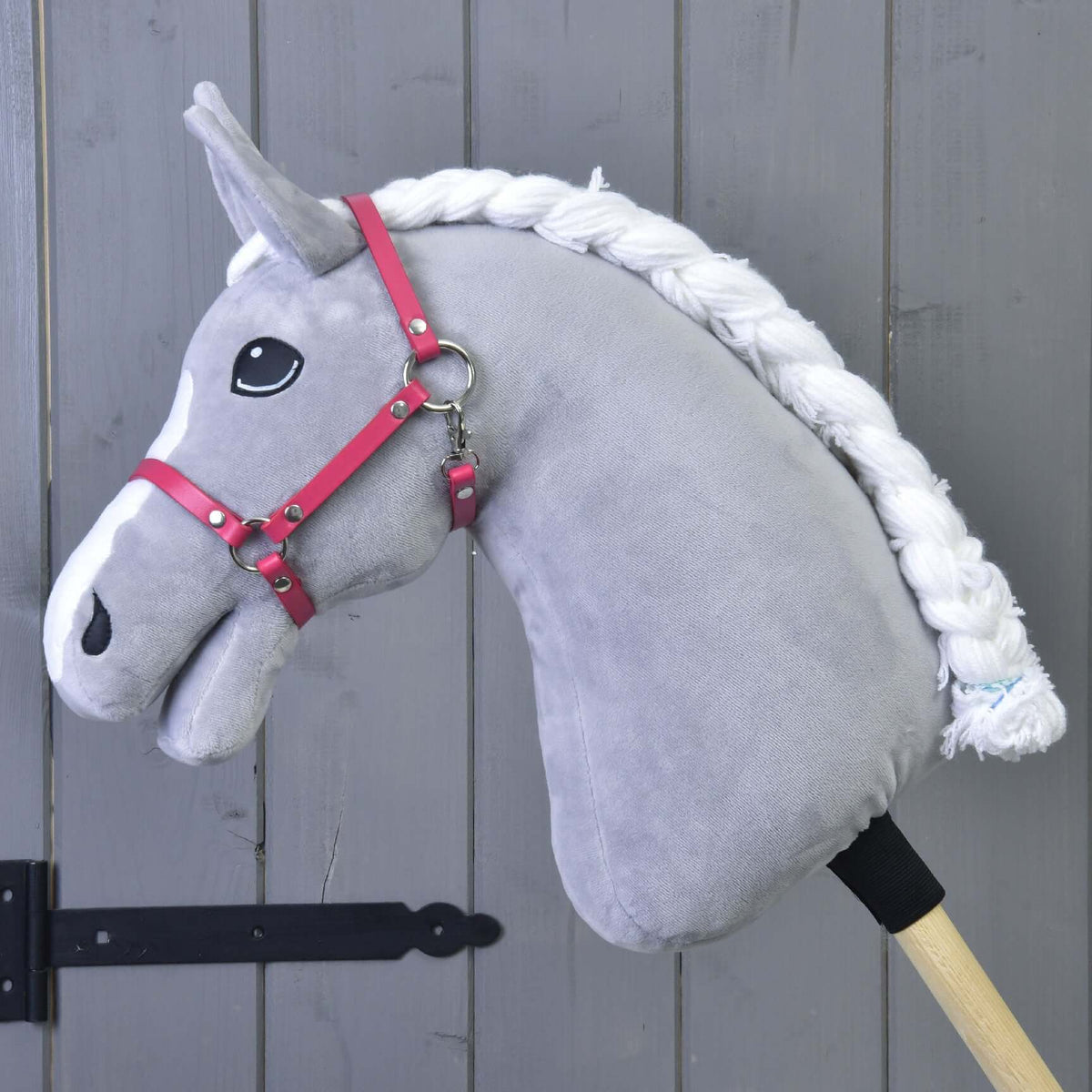 Szürke plüss Hobby Horse ló fehér hókával és fonott sörénnyel, a málnarózsaszín kötőfékben, szürke fafal előtt
