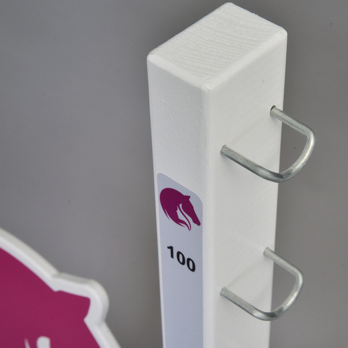 Közeli kép egy fehér oszlop tetejéről U alakú fém tartókampókkal és a 100-as jelzés fölötti kis rózsaszín lólogóval