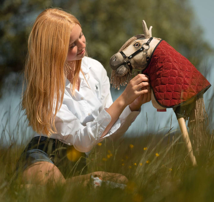 Girl in a white shirt sitting in a summer meadow, adjusting the red quilted blanket of her Hobby Horse