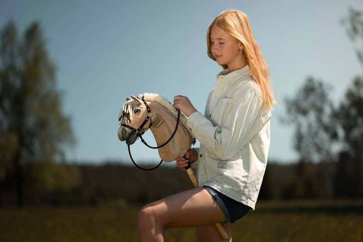 Girl in a light denim jacket holding a cream Hobby Horse with a black bridle and reins in a sunny meadow