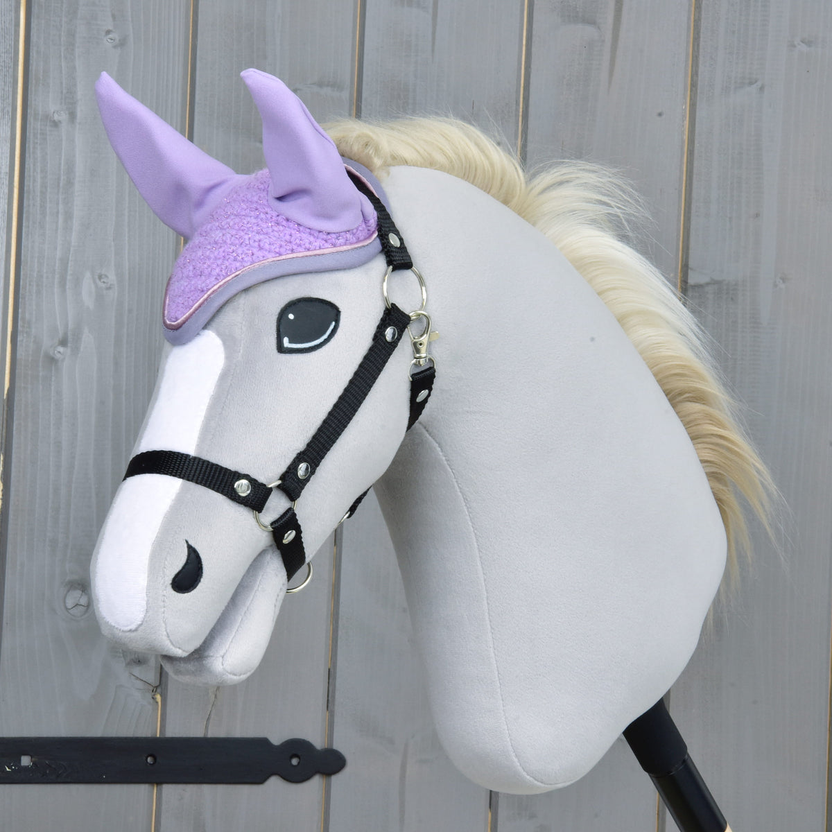 Világosszürke Hobby Horse ló fekete kötőfékkel és krémszínű sörénnyel, a lila horgolt fülvédőben