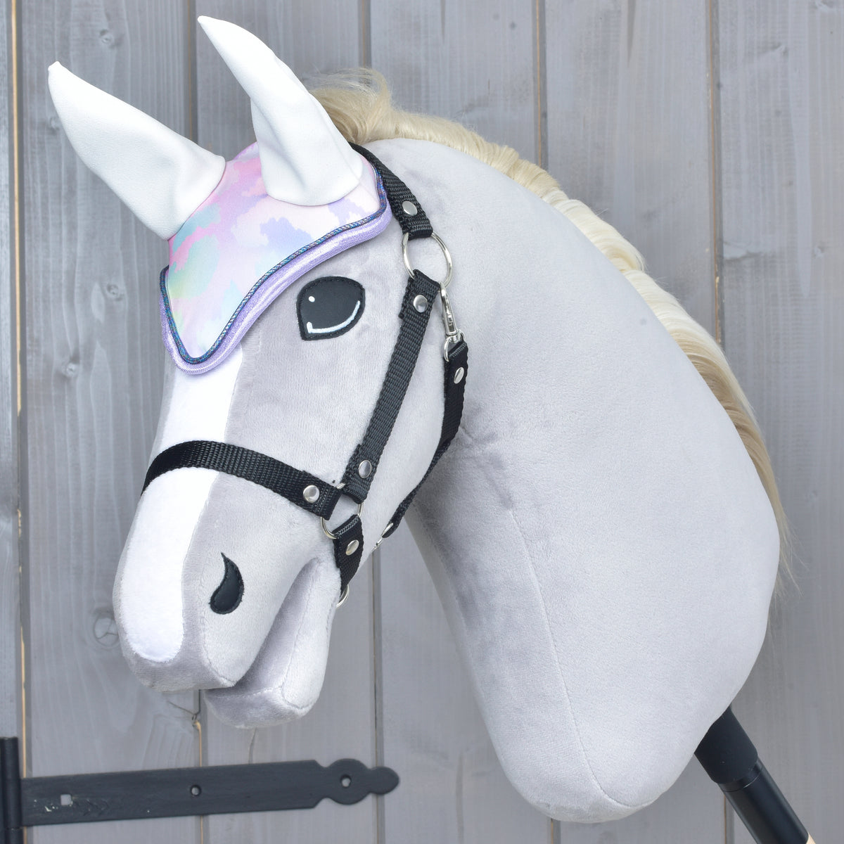 Light grey Hobby Horse wearing the pastel ear bonnet with white ears and a black halter against a wooden wall