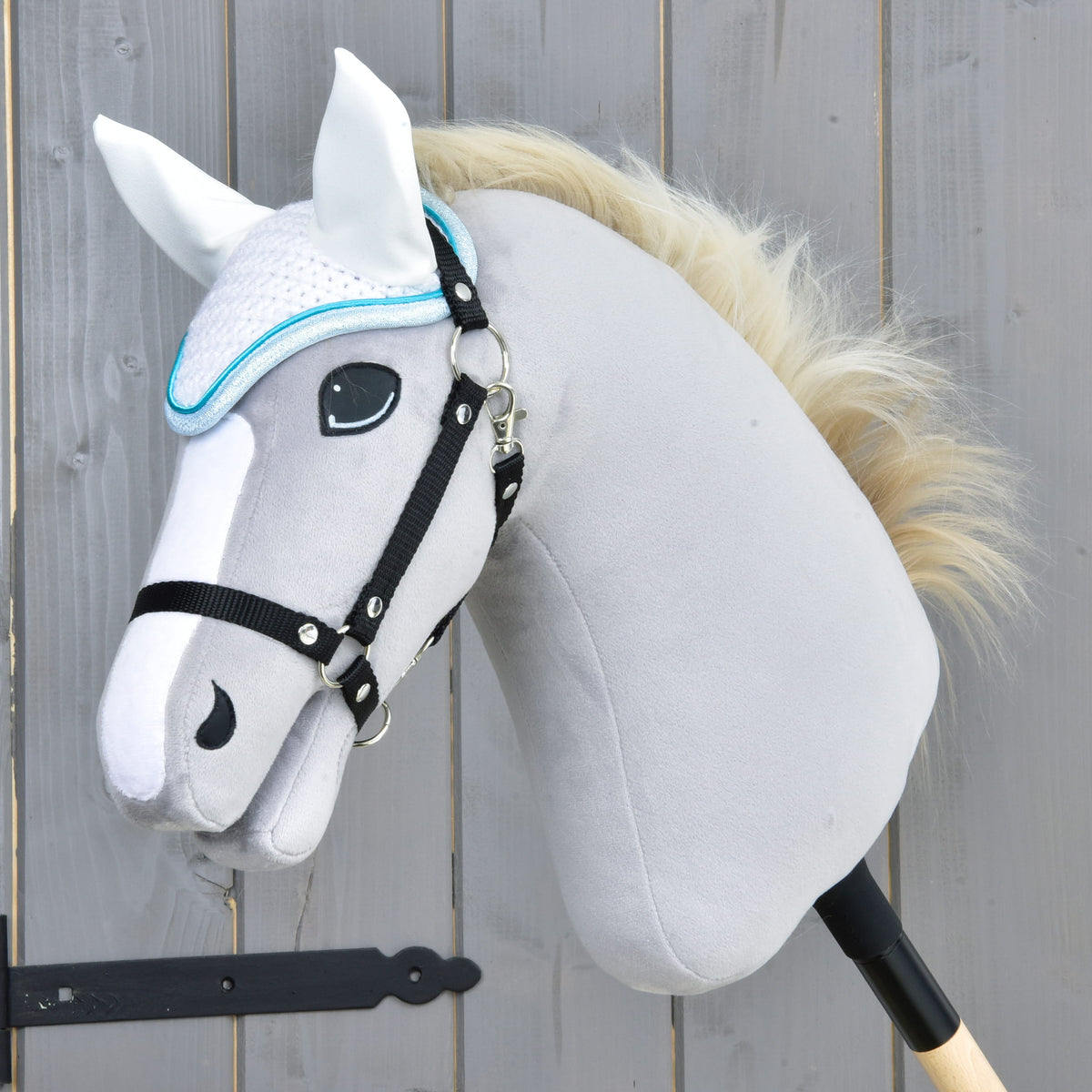 Világosszürke Hobby Horse ló fekete kötőfékkel és krémszínű sörénnyel, a türkiz szegélyű fehér horgolt fülvédőben