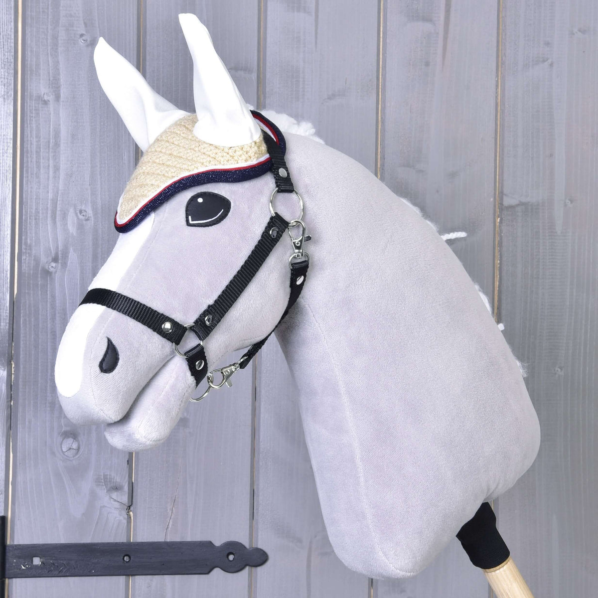 Szürke Hobby Horse ló fekete kötőfékben, a krémszínű horgolt Nicol fülvédőben fehér fülrészekkel és piros-sötétkék szegéllyel