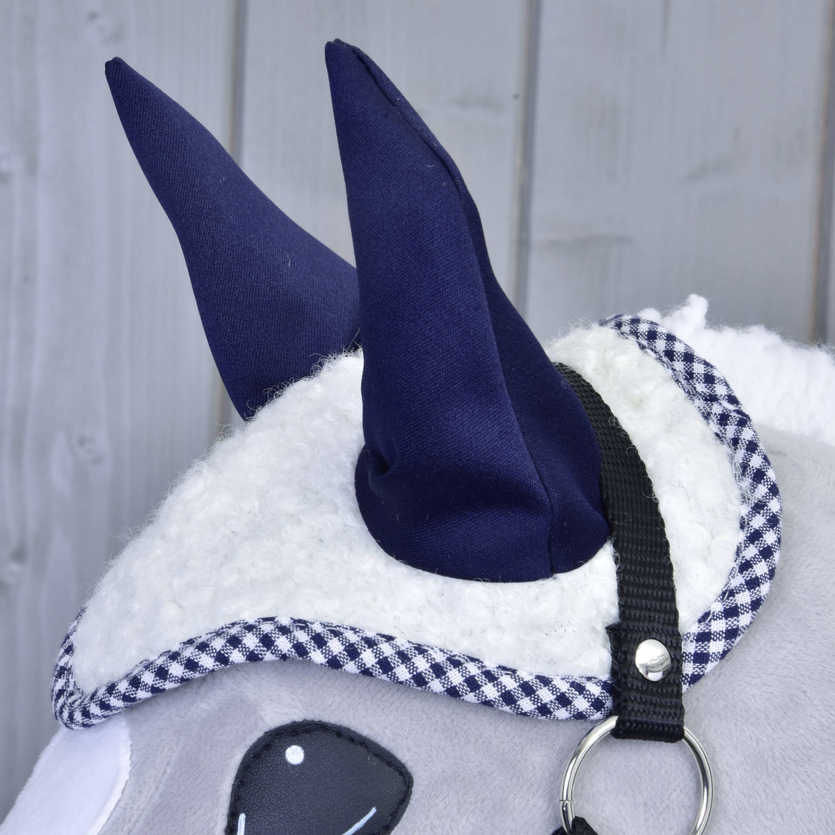 A fehér sherpa fülvédő közelről sötétkék fülekkel és vichykockás szegéllyel egy fekete kötőfékes szürke Hobby Horse lovon