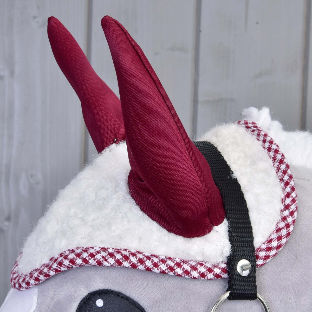 A fehér sherpa fülvédő bordó fülei és piros kockás szegélye egy szürke Hobby Horse lovon, látszik a fekete kötőfék szíja