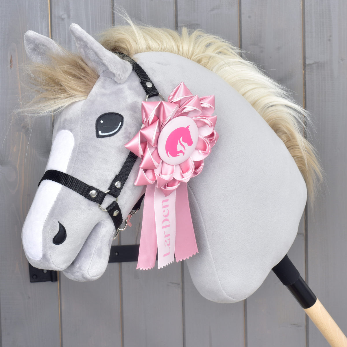 A LarDen Pink Hobby Horse rozetta egy fehér hókás, szőke sörényű szürke plüssfej fekete kötőfékére tűzve