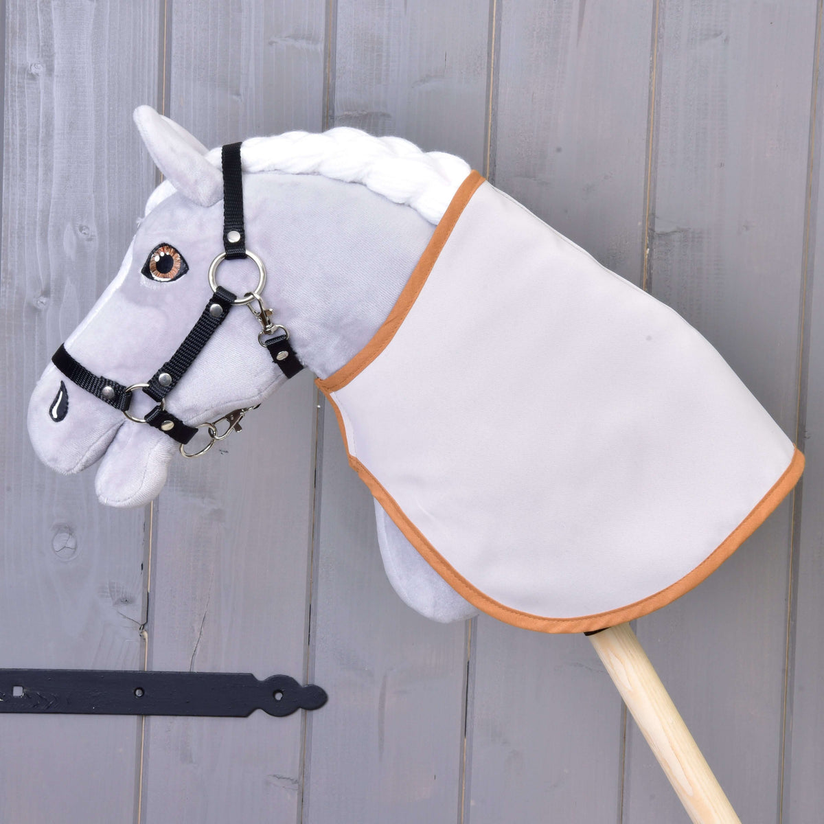 Szürke plüss Hobby Horse ló fekete kötőfékben, rajta a takaró az egyszínű világos oldalával kifelé, karamellszínű szegéllyel