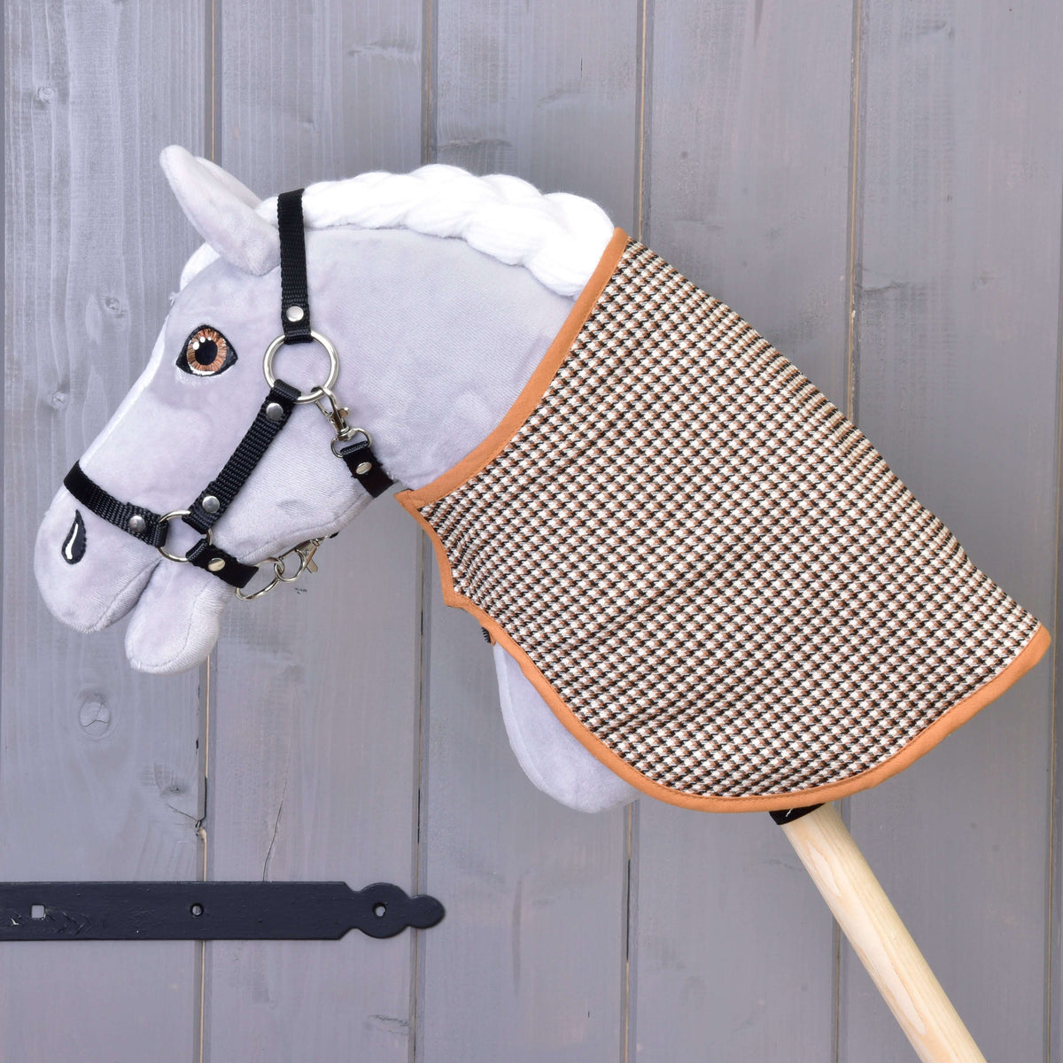 Szürke plüss Hobby Horse ló fekete kötőfékben, rajta a takaró a barna tyúklábmintás oldalával kifelé, szürke fal előtt