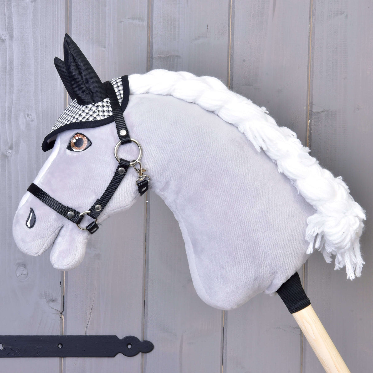 Szürke plüss Hobby Horse ló fonott fehér sörénnyel és fekete kötőfékkel, rajta a fekete-fehér tyúklábmintás fülvédő