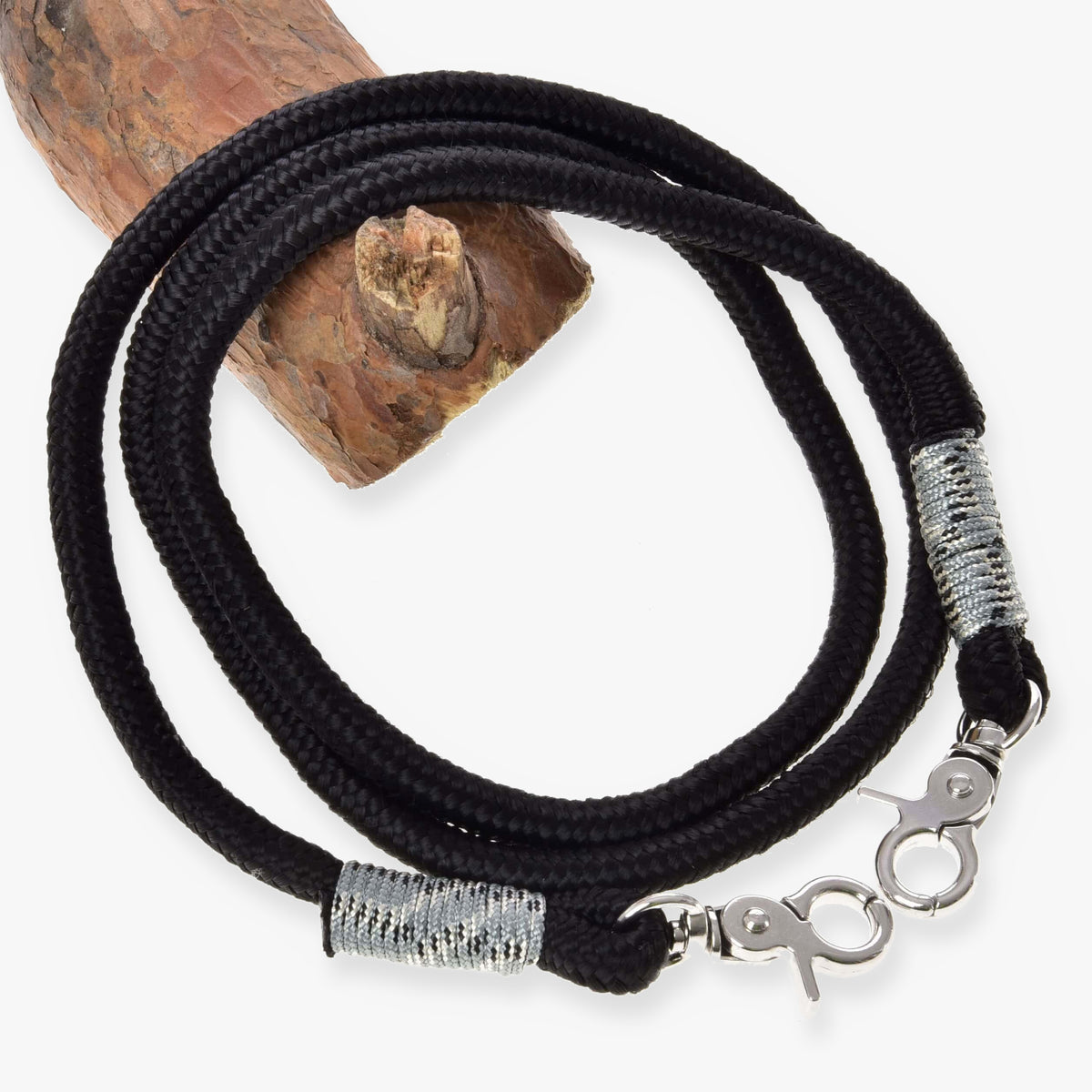 LarDen Hobby Horse Reins Black, braided black cord with silver-grey wraps and two silver snap hooks, coiled on a wooden log
