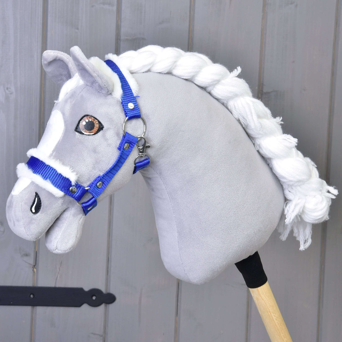 Szürke plüss Hobby Horse ló fonott fehér sörénnyel és barna szemekkel, rajta a királykék kötőfék fehér szőrmével
