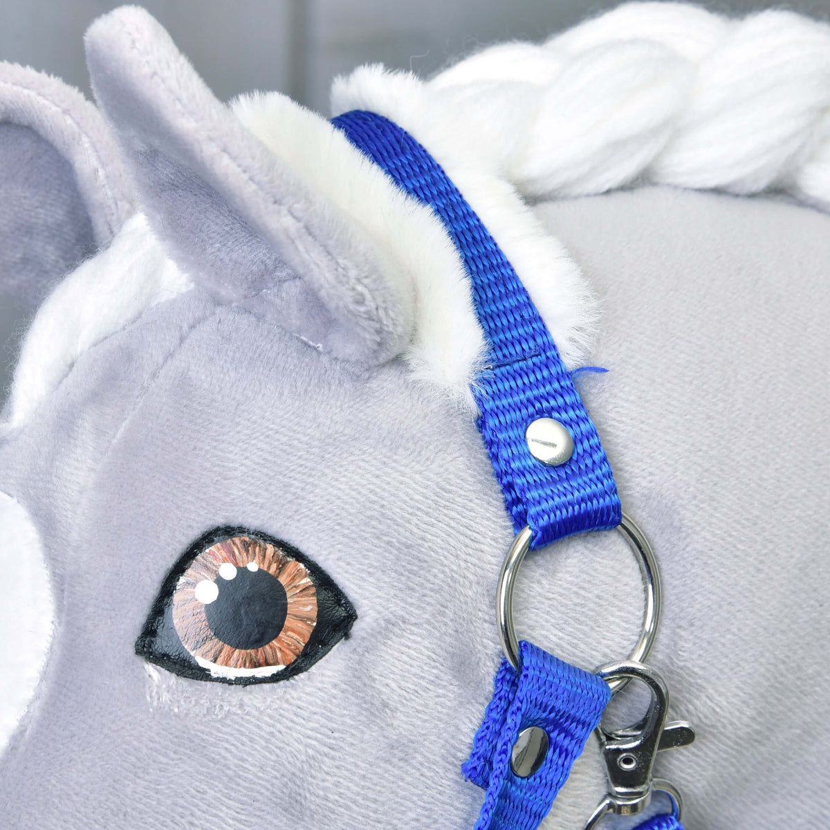 Közeli kép: a kék tarkószíj fehér szőrmével egy szürke Hobby Horse ló füle mögött, ezüstszínű karikával és karabinerrel