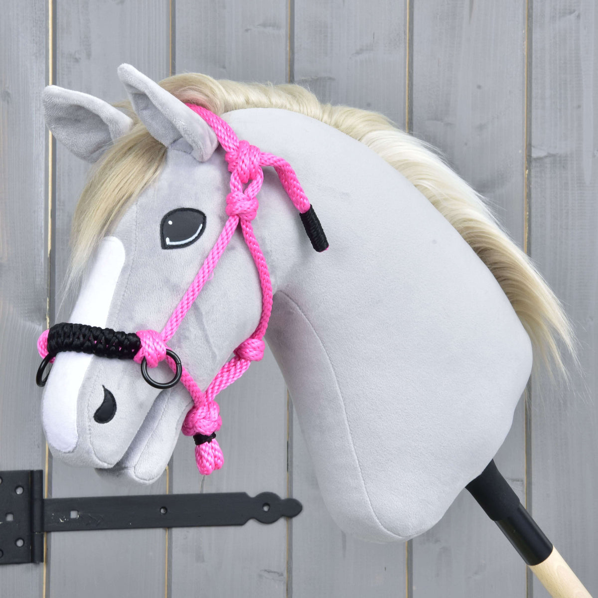 Light grey Hobby Horse with a blond mane wearing the pink rope halter with black noseband by a grey wooden door