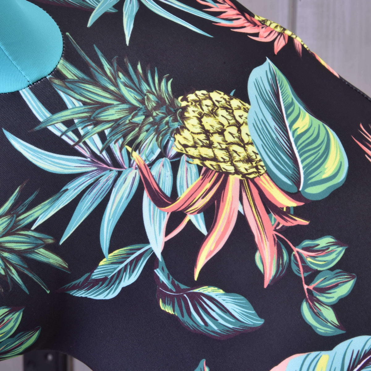A Pineapple Hobby Horse védőruha mintarészlete: sárga ananász, türkiz és korallszínű levelek fekete Lycra anyagon