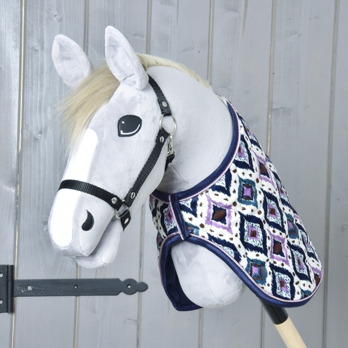 Krémszínű sörényű, fekete kötőfékes szürke Hobby Horse ló az Indian takaróban, elölnézetből egy szürke faajtónál