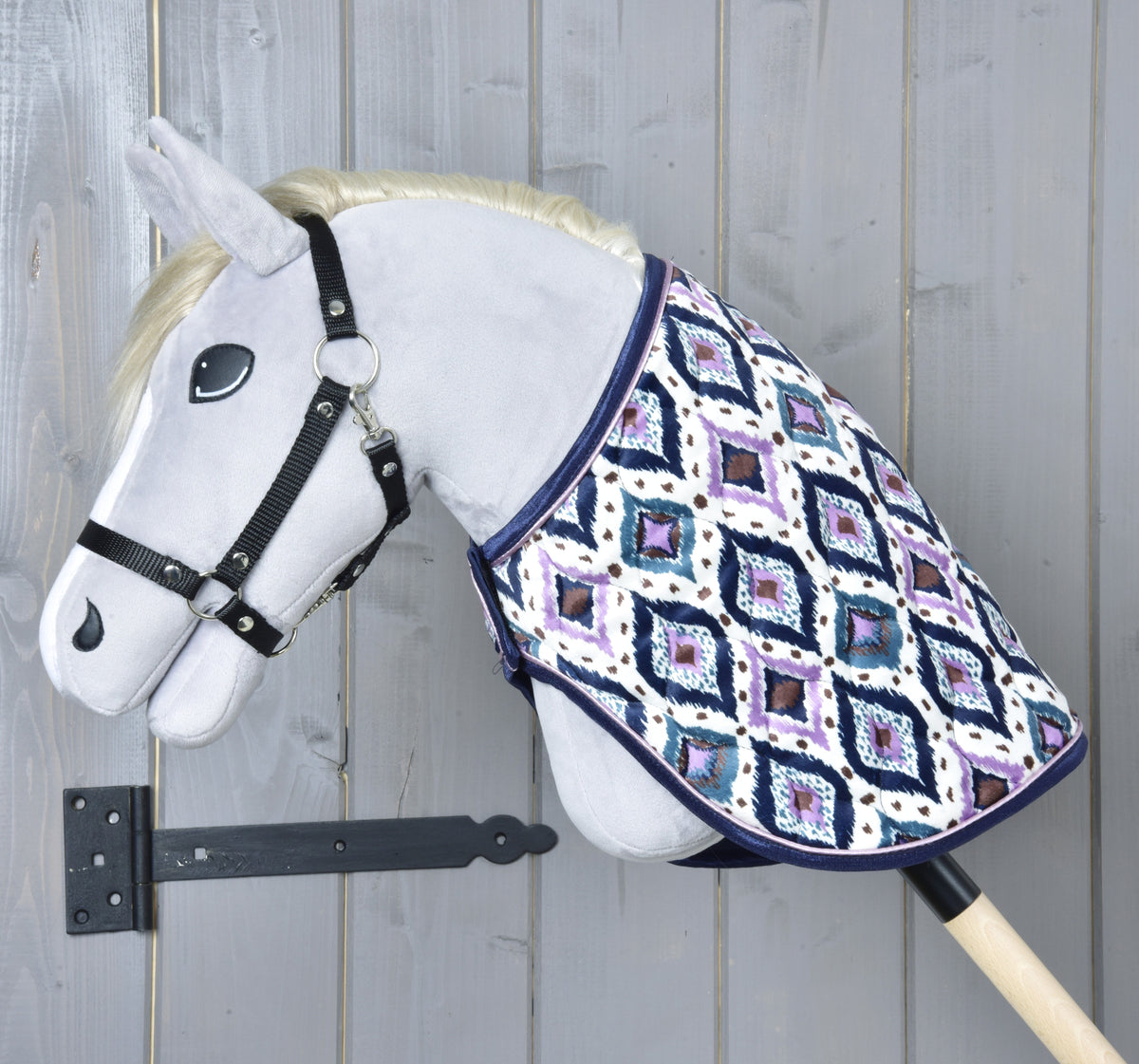 Fekete kötőfékes szürke Hobby Horse ló bal profilból, a nyakát az Indian takaró fedi, mögötte szürke fafal