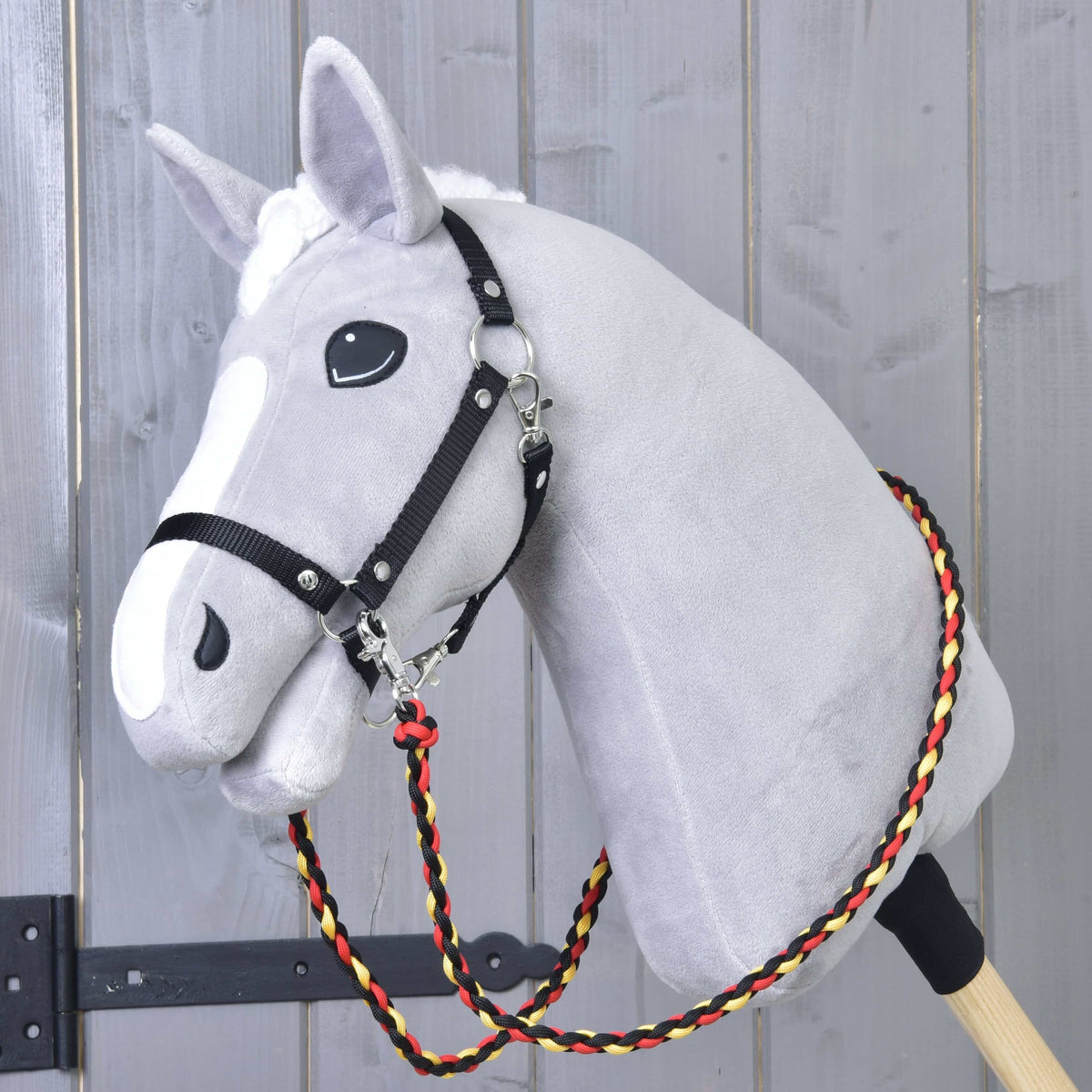Világosszürke Hobby Horse ló fekete kötőfékben, a nyaka körül a fekete-piros-sárga fonott Flag kantárszár