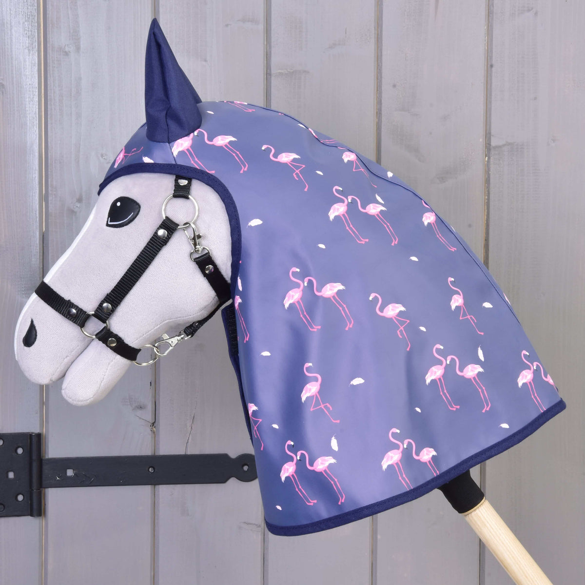 A flamingómintás esőtakaró oldalnézetből, amint egy szürke Hobby Horse ló fejét és nyakát takarja, szürke fafal mellett