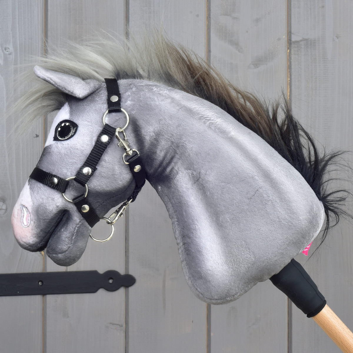 A fehér hókás szürke Hobby Horse ló oldalról, rózsaszín orral és fekete kötőfékkel, faboton