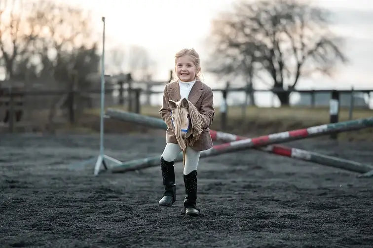 Tweedzakós, lovaglócsizmás kislány krémszínű Hobby Horse lóval játszik egy homokos pályán, akadályrudak között