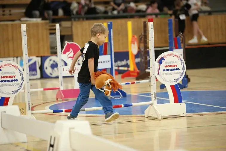 Kisfiú egy Hobby Horse versenyen, Hobby Horse lóval sétál a tornaterem padlóján, körülötte színes akadályok és Hobby Horsing rendezvénytáblák.