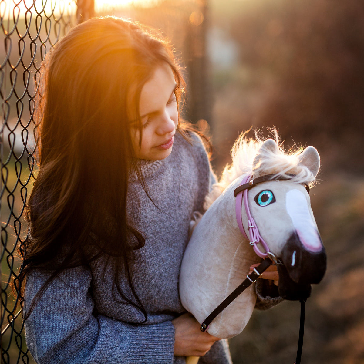Girl in a grey knitted sweater with a white Hobby Horse in a pink bridle at a fence in golden evening light