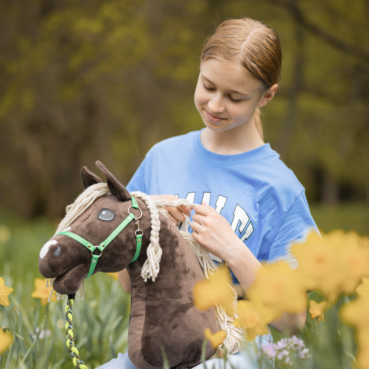 Kék pólós lány egy zöld kötőfékes, sötétbarna Hobby Horse ló sörényét fonja sárga nárciszok között