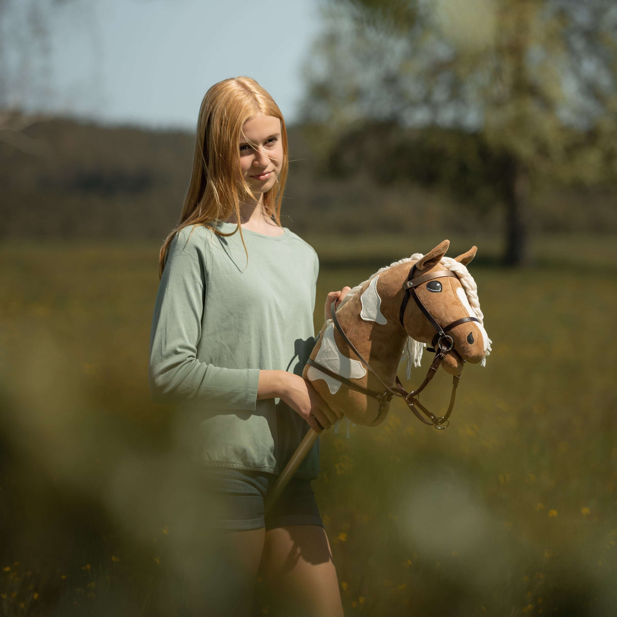 Girl in a sage green top in a meadow with a light brown pinto Hobby Horse in a brown bridle, reins and martingale