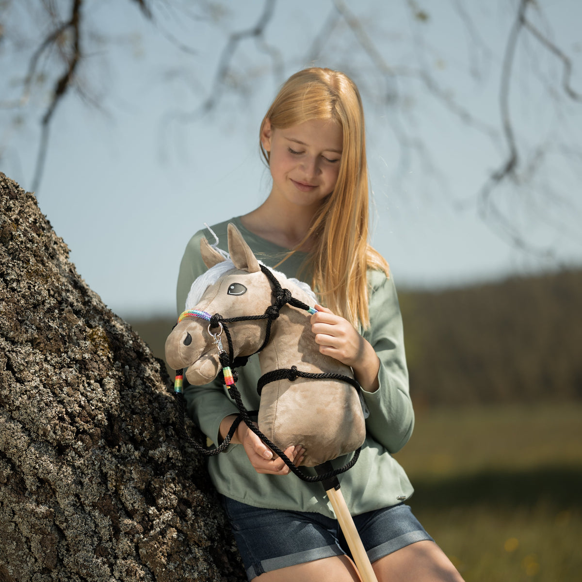 Blonde girl leaning on a tree trunk with a beige Hobby Horse in a black rope halter and reins with rainbow accents