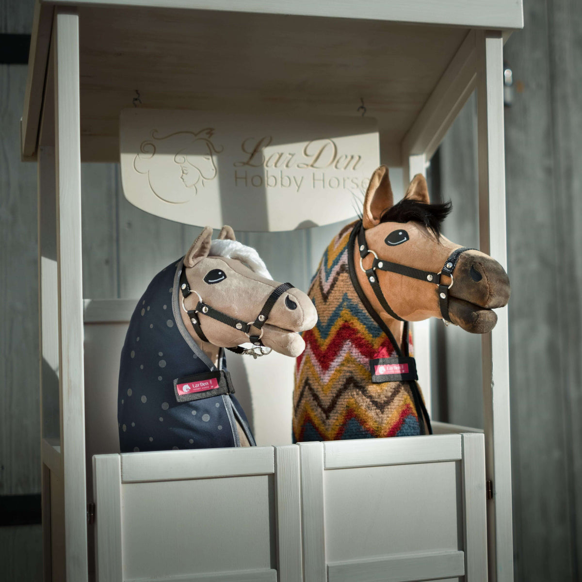 Two Hobby Horses in a white wooden stable box marked LarDen Hobby Horse, in a navy dotted and a colourful zigzag blanket